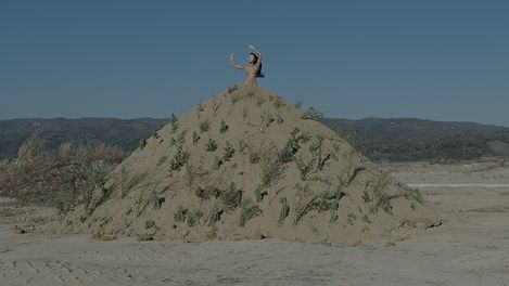 A person stands on a large sand mound with sparse vegetation raising one arm against a background of hills and blue sky.