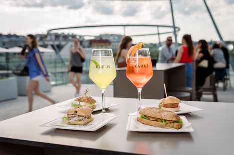 Two KATTUS drinks and several filled rolls on plates in an outdoor terrace area, with guests in the background.