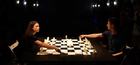 Two people sit opposite each other at a lit chessboard and move chess pieces.