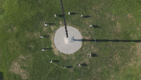 Aerial view of a group of people standing in a circle around a central object on a grassy area with long shadows cast by the sun