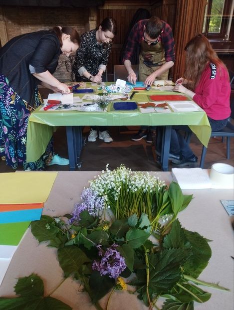 Four people stand around a table with craft materials working on a project, with various green leaves and flowers on another table in the foreground.