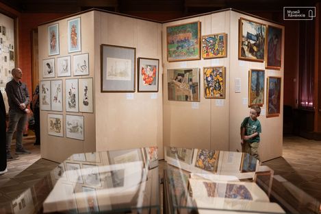 Visitors view paintings and drawings in a museum with light-colored walls and glass display cases in the foreground.