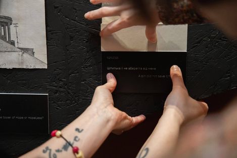 Hands holding a card with Cyrillic and English text against a black wall with carved lines.