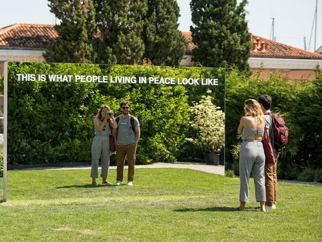 Zwei Personen stehen vor einem großen Spiegel im Freien, der den Text 'THIS IS WHAT PEOPLE LIVING IN PEACE LOOK LIKE' zeigt.