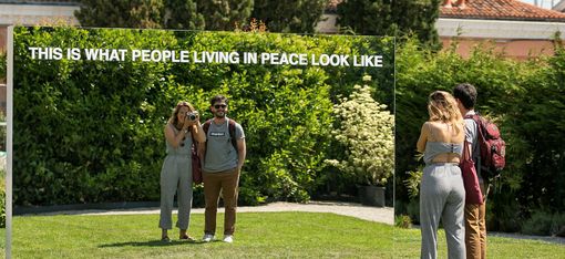 Zwei Personen stehen vor einem großen Spiegel im Freien, der den Text 'THIS IS WHAT PEOPLE LIVING IN PEACE LOOK LIKE' zeigt.
