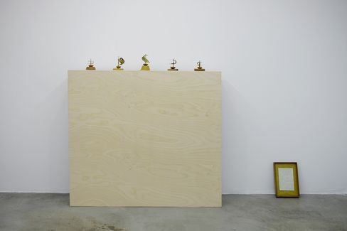 Five small sculptures on a large light wooden board against a white wall and a framed picture on the floor to the right.