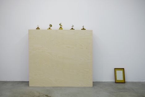 Five small sculptures on a large light wooden board against a white wall and a framed picture on the floor to the right.