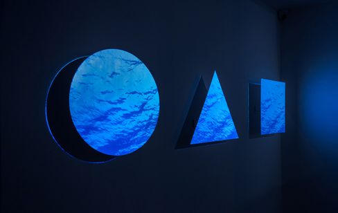Three metal shapes (circle, triangle, square) on a wall with blue video projection.