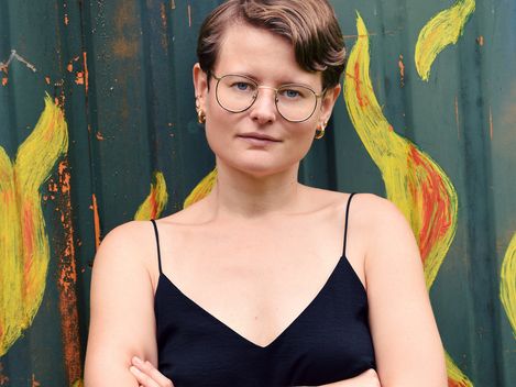 Person with short hair wearing a black sleeveless top and yellow pants standing with arms crossed in front of a wall painted with yellow and red flames.