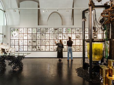 Zwei Personen betrachten eine Wand mit mehreren schwarz-weißen Zeichnungen in einem Ausstellungsraum mit großen Fenstern und technischen Installationen im Vordergrund