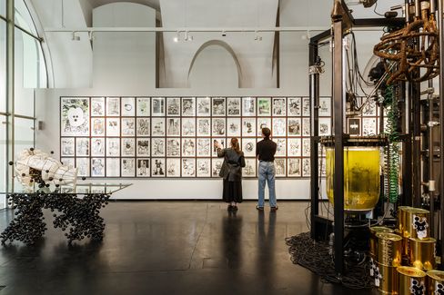 Two people looking at a wall of multiple black-and-white drawings in an exhibition space with large windows and technical installations in the foreground