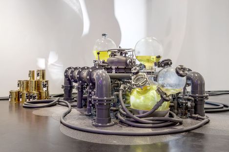 An intricate art installation with glass orbs, pipes, and yellow liquid, conveying an industrial and futuristic feel. Gold cylinders are stacked nearby.