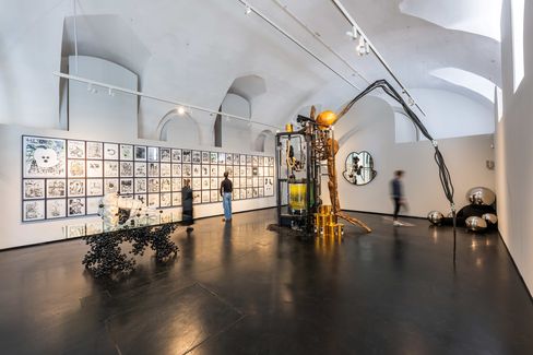 Exhibition view METABOLICA – Thomas Feuerstein at MQ Freiraum. Large exhibition space with white vaulted ceiling, a wall with many square drawings, several sculptures, and three people viewing the artworks.