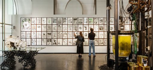 Zwei Personen betrachten eine Wand mit mehreren schwarz-weißen Zeichnungen in einem Ausstellungsraum mit großen Fenstern und technischen Installationen im Vordergrund