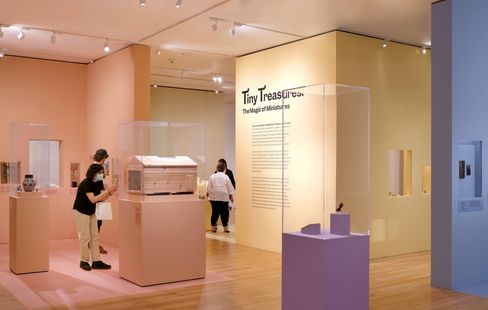 Museum exhibition with display cases and visitors in front of a wall reading "Tiny Treasures The Magic of Miniatures".