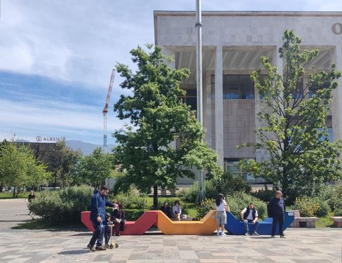 Mehrere Enzos in Rot, Gelb und Blau stehen auf einem gepflasterten Platz, auf denen Menschen sitzen und sich aufhalten, vor einem öffentlichen Gebäude mit Säulen und Bäumen in einer urbanen Umgebung.