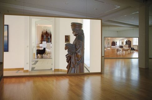 Large room with two adjacent screens each showing a museum scene with sheep and a statue