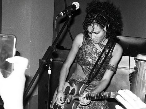 Woman with curly hair and headphones playing electric guitar on stage wearing patterned clothing and large boots.