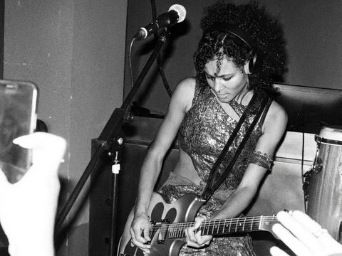Woman with curly hair and headphones playing electric guitar on stage wearing patterned clothing and large boots.