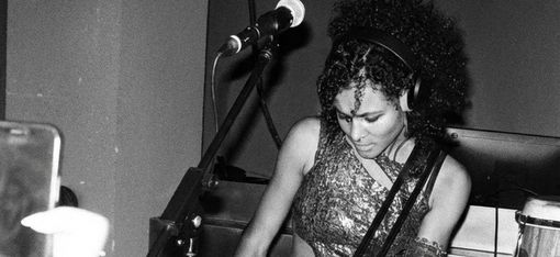 Woman with curly hair and headphones playing electric guitar on stage wearing patterned clothing and large boots.