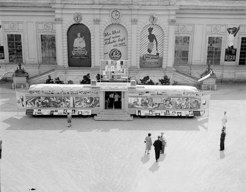 Schwarz-Weiß-Fotografie einer Wanderausstellung im Ehrenhof des Wiener Messepalasts in den 1950er-Jahren. Ein großer Ausstellungsbus mit Informations- und Werbetafeln steht vor der historischen Fassade, während Besucher:innen die Ausstellung besichtigen.