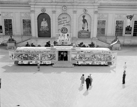 Schwarz-Weiß-Fotografie einer Wanderausstellung im Ehrenhof des Wiener Messepalasts in den 1950er-Jahren. Ein großer Ausstellungsbus mit Informations- und Werbetafeln steht vor der historischen Fassade, während Besucher:innen die Ausstellung besichtigen.