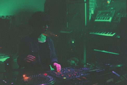 Person at DJ console with mixer and turntables in dark room with green lighting.
