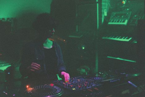 Person at DJ console with mixer and turntables in dark room with green lighting.