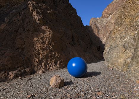 Large blue sphere on gravelly ground between tall rocky cliffs under clear blue sky.