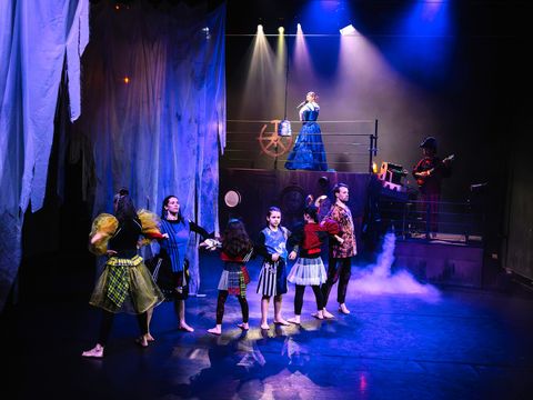 Six people of different ages form a strong chain with their arms, demonstrating their strength. In the background, a singer in a blue dress and a musician stand on a ship.