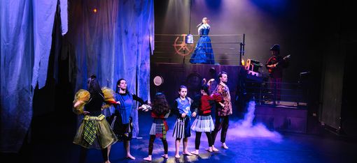 Six people of different ages form a strong chain with their arms, demonstrating their strength. In the background, a singer in a blue dress and a musician stand on a ship.