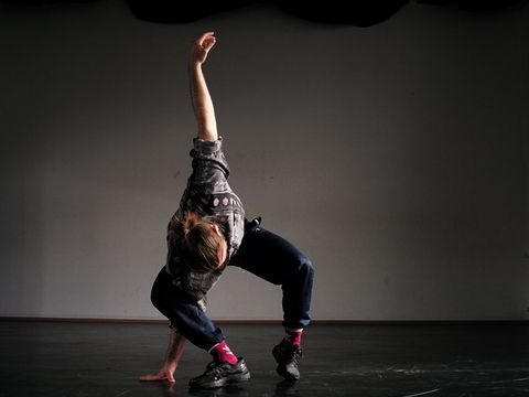 Person in casual clothing and pink socks in a dynamic dance pose on a dark floor against a gray wall