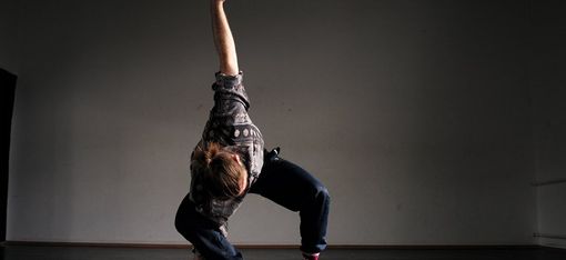 Person in casual clothing and pink socks in a dynamic dance pose on a dark floor against a gray wall