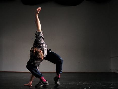 Person in lockerer Kleidung und pinken Socken in einer dynamischen Tanzpose auf einem dunklen Boden vor einer grauen Wand