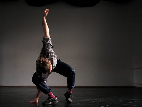 Person in lockerer Kleidung und pinken Socken in einer dynamischen Tanzpose auf einem dunklen Boden vor einer grauen Wand