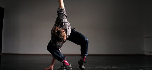 Person in casual clothing and pink socks in a dynamic dance pose on a dark floor against a gray wall