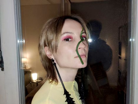 Person with short hair wearing a light, sheer top stands in front of a glass door reflecting another person in the background.