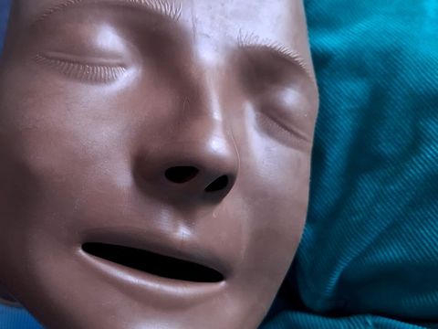 Nahaufnahme einer braunen Gesichtsmaske mit geschlossenen Augen und leicht geöffnetem Mund vor einem türkisfarbenen Hintergrund.