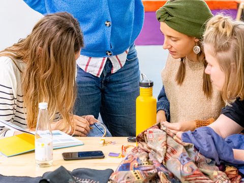 Drei Frauen sitzen an einem Tisch mit Stoffen und Nähmaterialien, eine vierte Frau steht und schaut auf die Arbeit am Tisch.