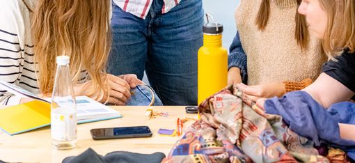 Drei Frauen sitzen an einem Tisch mit Stoffen und Nähmaterialien, eine vierte Frau steht und schaut auf die Arbeit am Tisch.