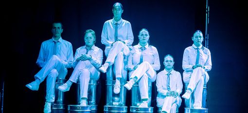 Six people are sitting on stacked metal buckets, dressed in white costumes.