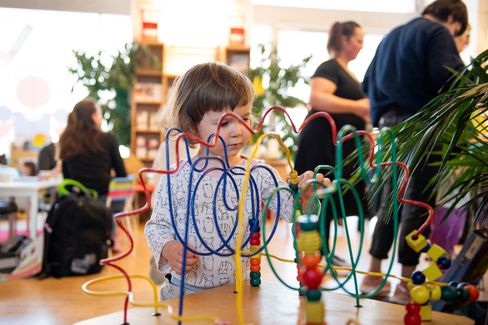 The image shows a small child playing intently with a bead maze toy made of colorful, curved wires and wooden beads. The child is standing at a low table and moving a bead along one of the wire tracks.

In the background, there is a bright, welcoming indoor space that looks like a family-friendly information or play area. You can see plants, shelves with materials, and several adults talking or զբաղված with different activities. One person is sitting on the floor, and a stroller is visible nearby.

The atmosphere feels lively and cozy – a place where children can play while adults talk, browse, or get advice.
