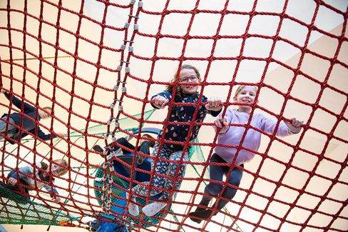 The image shows two children standing on a red climbing net in an indoor play area, smiling down at the camera. They are holding onto the net with their hands and appear happy and energetic. One of the children is wearing glasses and a dark blue patterned top, while the other is wearing a purple sweater.

Below them, you can see more parts of the rope and net climbing structure, as well as another child playing in the background. The space is bright and welcoming, resembling a gym or an indoor playground.

The photo is taken from a low angle, looking up, which makes the net and the children appear dynamic and gives a strong sense of height and movement.