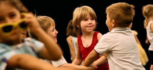 Zu sehen ist eine Gruppe von Kindern beim Spielen, im Vordergrund unscharf ein Kind, das gerade eine Sonnenbrille abnimmt und offensichtlich in Bewegung ist, daneben etwas weiter hinten eine Dreiergruppe, die sich an den Händen hält und gerade miteinander spricht, im Hintergrund weitere Kinder, die nur zum Teil zu sehen sind von hinten oder von der Seite.