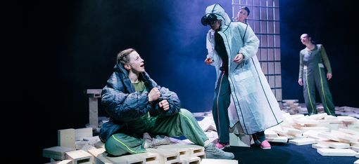 Das Bild zeigt eine Gruppe von drei Personen auf einer Bühne, die ein Theaterstück aufführen. Eine Person steht auf der linken Seite der Bühne, während die anderen beiden auf der rechten Seite stehen. Sie scheinen miteinander zu interagieren, möglicherweise diskutieren sie oder proben ihre Darbietung. Die Bühne hat eine einzigartige Kulisse, mit zahlreichen Blöcken, die über den Bereich verstreut sind. Einige dieser Blöcke sind in der Nähe der Personen platziert, während andere weiter entfernt stehen. Die Gesamtatmosphäre der Szene suggeriert eine kreative und fesselnde Darbietung.