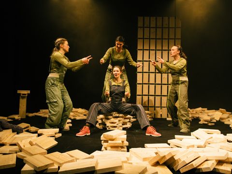 The picture shows a group of four women standing on a stage, each wearing green outfits. They are striking different poses: one woman is sitting on a pile of wooden blocks, whilst the others are standing around her. The women are in the middle of a performance and are interacting with one another using sign language. Several wooden blocks are scattered across the stage, some of which are close to the women, whilst others are further away. The scene captures the dynamic energy of the performance.
