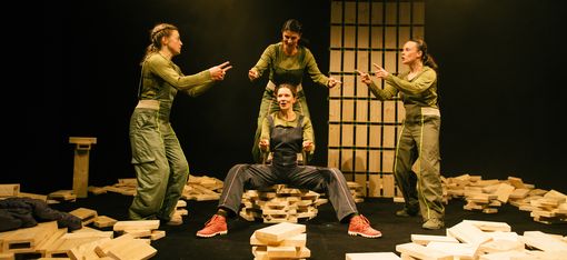 The picture shows a group of four women standing on a stage, each wearing green outfits. They are striking different poses: one woman is sitting on a pile of wooden blocks, whilst the others are standing around her. The women are in the middle of a performance and are interacting with one another using sign language. Several wooden blocks are scattered across the stage, some of which are close to the women, whilst others are further away. The scene captures the dynamic energy of the performance.