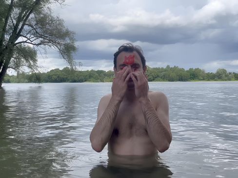 A man stands waist-deep in a lake or river water with his hands covering his face and a red symbol on his forehead.