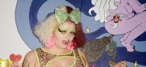 Person mit zweifarbiger Frisur, grünem Haarband, auffälligem Make-up und Netzoberteil vor einer bunten Wand mit einer gemalten Figur.