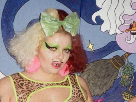 Person with two-tone hair, green hair bow, bold makeup, and fishnet top in front of a colorful wall with a painted figure.